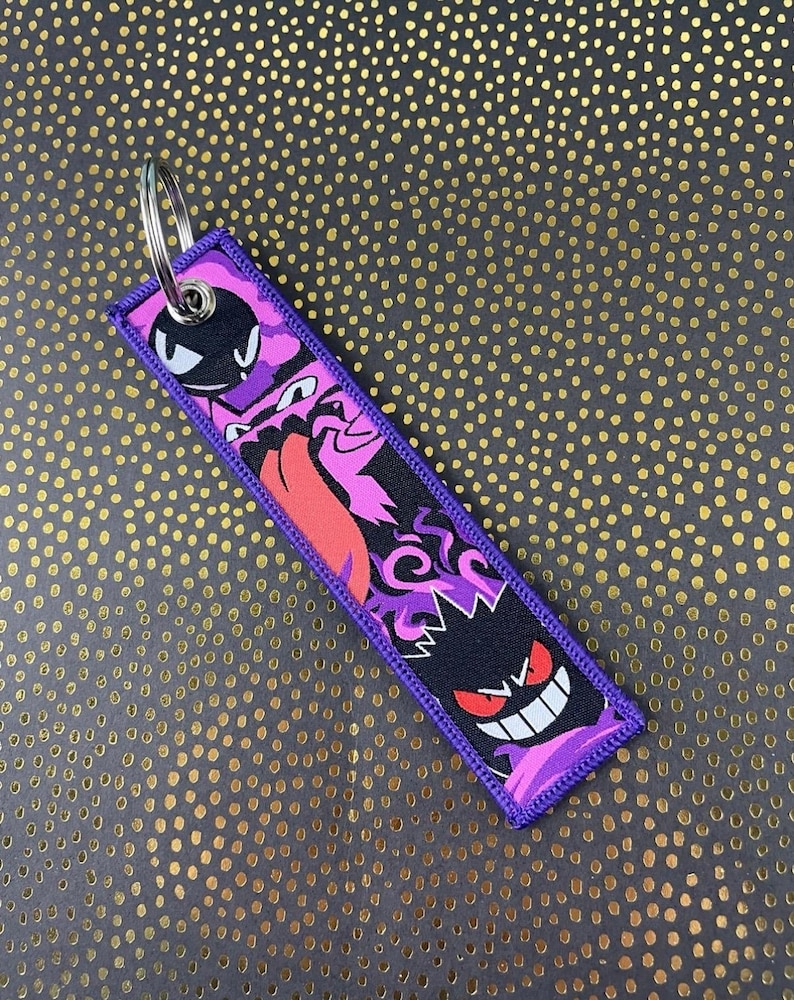 May include: A rectangular purple keychain featuring a design of cartoon ghosts in black, pink, and red. The keychain has a silver metal ring for attaching keys. The edges are stitched with purple thread.