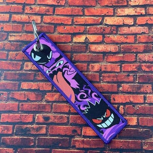 May include: A rectangular purple and black keychain featuring cartoon ghost characters. The keychain has a purple border and a silver key ring. The background is a red brick wall.