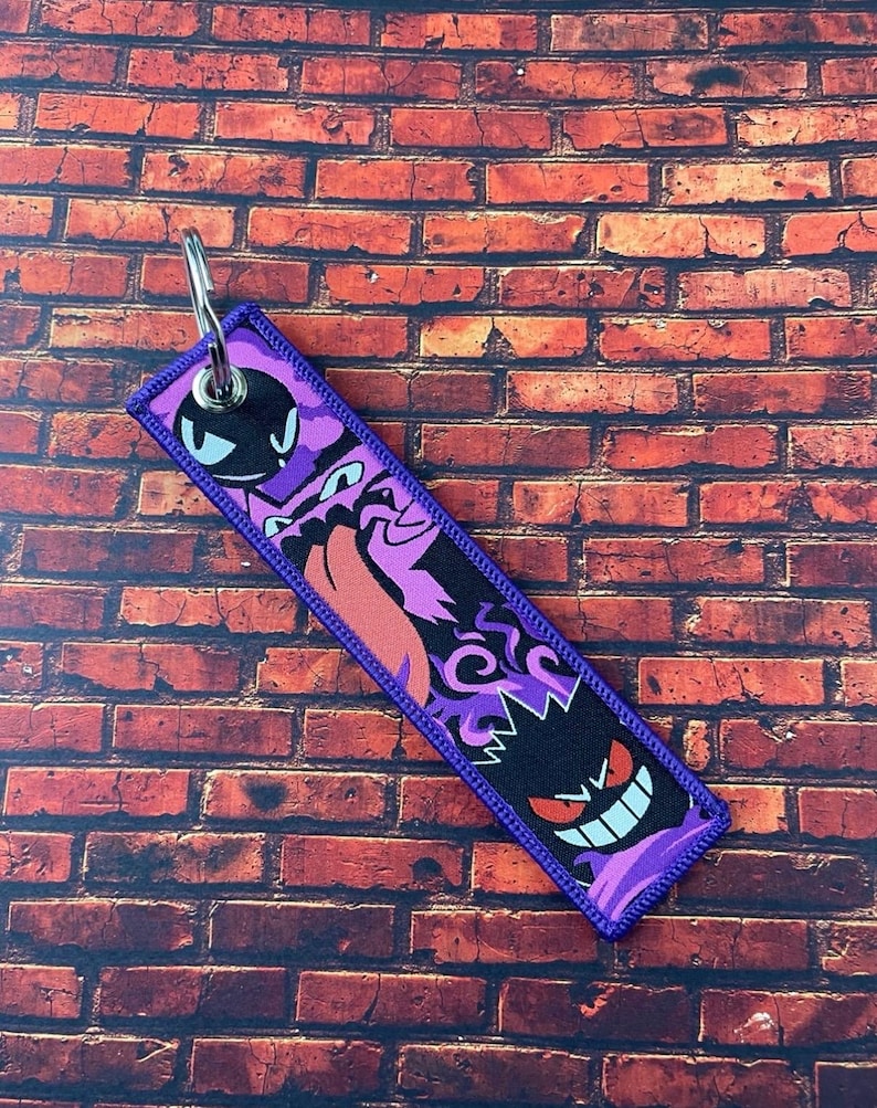 May include: A rectangular purple and black keychain featuring cartoon ghost characters. The keychain has a purple border and a silver key ring. The background is a red brick wall.