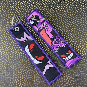 May include: Two black keychains with purple trim and silver key rings. Each keychain features a different cartoon ghost character in shades of purple, pink, red, and white. The background is a gold and black dotted pattern.