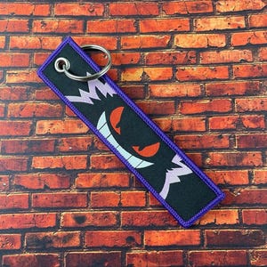 May include: Black rectangular keychain with a purple border and a silver key ring. The keychain features a cartoon ghost face with red eyes, a white smile, and purple accents.