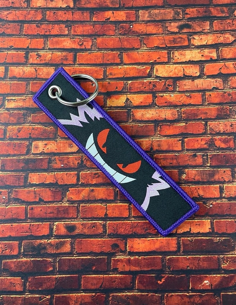 May include: Black rectangular keychain with a purple border and a silver key ring. The keychain features a cartoon ghost face with red eyes, a white smile, and purple accents.
