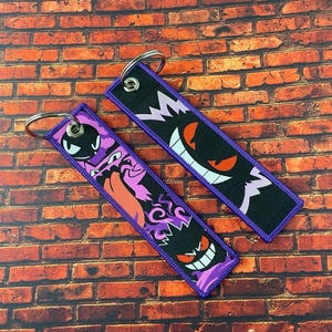 May include: Two rectangular keychains with embroidered designs. One features a purple and black ghost with a menacing grin, the other a similar ghost with a different expression. Both have purple borders and silver key rings.