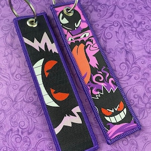 May include: Two rectangular keychains with purple trim and silver key rings. One keychain features a black design with a white and pink lightning bolt and a red-eyed ghost. The other has a pink and purple design with a cartoon ghost.