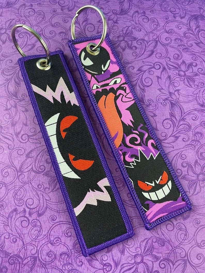 May include: Two rectangular keychains with purple trim and silver key rings. One keychain features a black design with a white and pink lightning bolt and a red-eyed ghost. The other has a pink and purple design with a cartoon ghost.