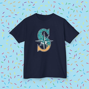 May include: Navy blue t-shirt featuring a stylized "S" logo with a baseball theme. The "S" is decorated with blue sprinkles and a waffle cone, with a compass rose in the center. The shirt is a classic crew neck.