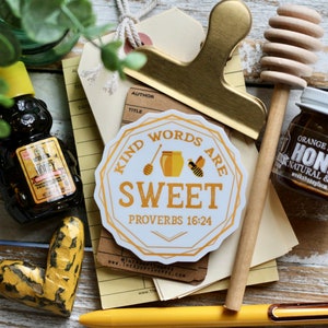 Könnte beinhalten: Ein weißer Aufkleber mit goldener Umrandung und einer Bienenillustration. Der Aufkleber lautet "Kind words are sweet" und "Proverbs 16:24".