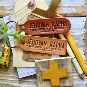 Può includere: Due portachiavi in ​​cuoio marrone con il testo "Jehovah Rapha + The Lord is my Healer + Jeremiah 17:14" in rilievo. I portachiavi sono su una superficie di legno con un cuore giallo e una piccola croce di legno.