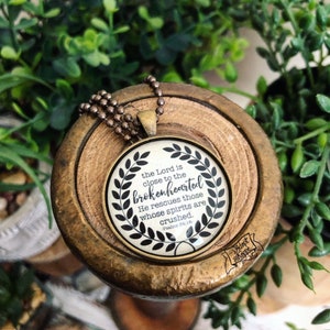 May include: A bronze necklace with a round glass pendant featuring a black and white design with the text "the Lord is close to the brokenhearted He rescues those whose spirits are crushed. Psalms 34:18"