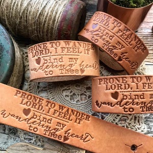 May include: Three leather bracelets with the text "Prone to wander Lord, I feel it bind my wandering heart to Thee" engraved on them. The bracelets are brown and have a heart symbol engraved on each one.