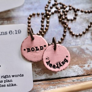 May include: A pink necklace with two round pendants. The pendants have the words "BOLD" and "healing" inscribed on them. The necklace is made of a brass chain.
