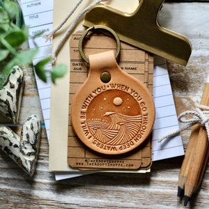 Puede incluir: Llavero de cuero marrón con el texto "Estaré contigo - cuando pases por aguas profundas" y el versículo bíblico "Isaías 43:2". El llavero presenta un diseño de olas y una luna.