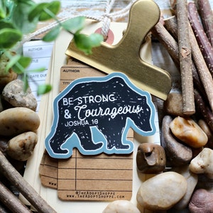 May include: A black bear sticker with a blue outline. The sticker has the text "Be Strong & Courageous Joshua 19" on it. The sticker is on a brown surface with rocks and sticks.