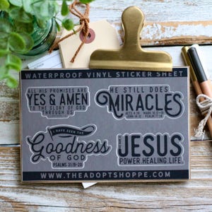 Pode incluir: Uma folha de autocolantes de vinil impermeável cinzenta com quatro desenhos em branco e preto. Os autocolantes apresentam frases inspiradoras como "Yes & Amen" e "Miracles". A folha é segurada por um clip dourado, com lápis e uma etiqueta no fundo.