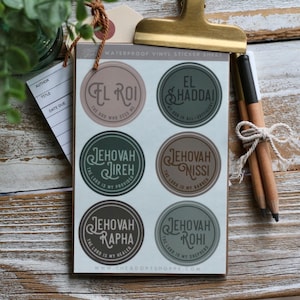 May include: A sheet of waterproof vinyl stickers featuring six circular designs with religious phrases. Each sticker has a different color scheme and text, including "El Roi" and "Jehovah Jireh". The stickers are held by a gold clip.