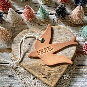 May include: A rustic wooden heart-shaped sign with a leather bird cutout that says "FREE." The bird is attached to the sign with a twine string. The sign is surrounded by small, colorful, fuzzy trees.