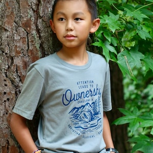 Pode incluir: Um menino jovem vestindo uma camiseta cinza claro com um gráfico azul que diz "Atenção leva à propriedade. Desafio! Um acampamento clássico cristão." Ele está em pé na frente de uma árvore e usando várias pulseiras coloridas no pulso.