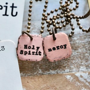 May include: Two pink square pendants on a gold chain necklace. The pendants have the words "Holy Spirit" and "mercy" inscribed on them.