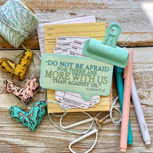 May include: A teal blue speech bubble with the text "Do not be afraid for there are more with us than against us." -2 Kings 6:16. The speech bubble is clipped to a yellow lined notepad with a teal blue binder clip. There are three wooden hearts in the image, one yellow, one pink, and one teal blue.