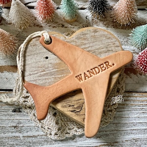 May include: A tan leather airplane ornament with the word "WANDER" embossed on the wing. The ornament is attached to a heart-shaped wooden base with a cream-colored string. Decorative miniature Christmas trees are in the background.