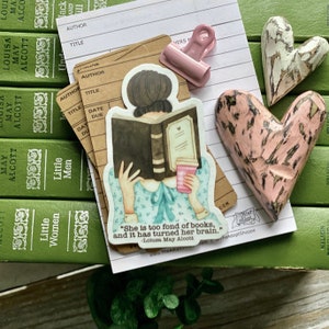 Pode incluir: Um adesivo com uma ilustração de uma mulher lendo um livro com uma xícara de café na outra mão. O adesivo tem uma citaat de Louisa May Alcott que diz: "Ela é muito fã de livros, e isso a deixou louca." O adesivo está colocado em uma pilha de livros com os títulos "Little Women" e "Little Men".