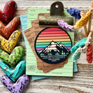 May include: A rainbow mountain sticker with the text "When my heart is overwhelmed, lead me to the rock that is higher than I."  The sticker is on a green and brown notepad with a clipboard.