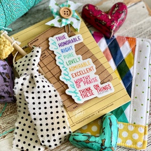 May include: A collection of decorative items including a sticker with the words "True, Honorable, Right, Pure, Lovely, Admirable, Excellent, Worthy of Praise" and the text "Think on these things". Also, a polka dot ribbon, a heart-shaped object, and a small fabric pinwheel.