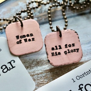 May include: Two pink square pendants on a gold chain necklace. The pendants have the text "Women of War" and "all for His glory" on them. The pendants are hanging over two cards with the text "189:15-18 of War" and "1 Corinthians 10:3 for"