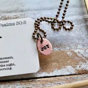 May include: A copper necklace with a round pendant that says "JOY" on it. The necklace is on a white card with the text "Psalms 30:5" and a quote about joy.