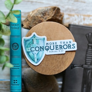 May include: A teal blue sticker with a watercolor shield and the text "More Than Conquerors Romans 8:37" on a wooden surface.