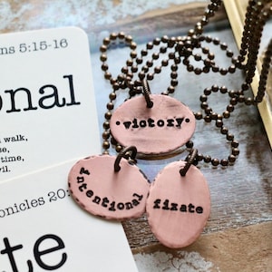 May include: A necklace with three oval-shaped pendants that say "victory", "intentional", and "fixate". The pendants are a light pink color and have a distressed look. The necklace is made of a brass chain.