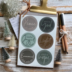 Pode incluir: Uma folha de adesivos de vinil à prova d'água com designs circulares e frases religiosas como "Paz na Terra" e "Glória a Deus". Os adesivos são em cores suaves e são presos por um clipe dourado. Também estão presentes pequenas árvores de Natal e lápis.