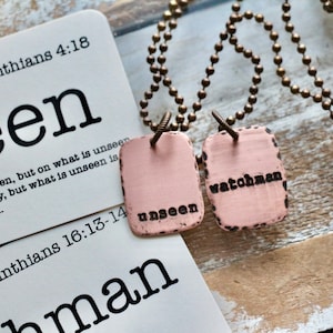 May include: Two pink metal tags on a brass chain necklace. The tags are rectangular and have the words "unseen" and "watchman" engraved on them. The necklace is accompanied by two cards with bible verses from 2 Corinthians 4:18 and 1 Corinthians 16:13-14.
