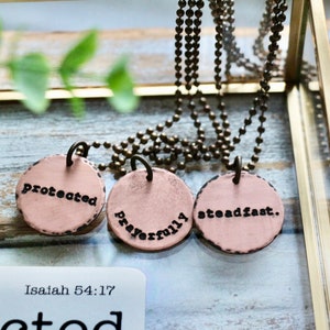 May include: Three round pink metal pendants on gold chains. The pendants have the words "protected", "prayerfully", and "steadfast" inscribed on them.