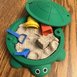 May include: A green turtle-shaped sandbox with a lid. The sandbox is filled with sand and includes a yellow bucket, a red brick, and two blue shovels.