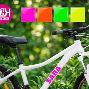 May include: White mountain bike with black tires and a black seat. The bike has the name "SARA" in pink lettering on the frame. The image also includes a logo and four colorful squares.