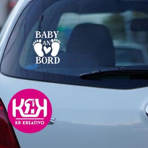 Op de afbeelding: Witte autosticker met de tekst "BABY AN BORD" boven een hart en voetstappen. Een roze cirkel met witte tekst en een logo staat eronder.