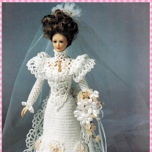 Puede incluir: Una muñeca de porcelana con un vestido de novia de ganchillo blanco de manga larga, cuello alto y falda con ribete de encaje. La muñeca tiene el pelo castaño recogido, un velo blanco transparente y sostiene un ramo de flores blancas.
