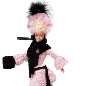May include: A doll with blonde hair wearing a pink crocheted gown with black trim, a matching pink bonnet with a black feather, a black scarf, and black cuffs. A small koala pin is attached to the gown. Text on the skirt reads: Compiled and Digitized by Aussie Sewing & Craft.