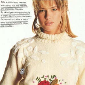 May include: Cream-colored sweater with a cable-knit neckline and floral embroidery. The front features a vibrant bouquet of red, orange, and purple flowers. White leaf patterns adorn the shoulders and edges. The text on the image says: Take a plain cream sweater with cabled ribs and neckline, and embroider it lavishly. An extravagant bouquet worked in bright tapestry yarns dominates the centre front, while a trail of white leaves frames the edges and shoulders.