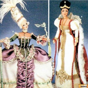 May include: Two dolls wearing elaborate historical-style costumes. The left doll features a tall white wig with feathers, a green and purple gown with lace trim, and a handheld mask. The right doll wears a white gown with gold trim and a red cape.
