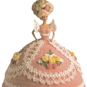 May include: A doll with blonde hair and a pink dress. The dress is crocheted with white lace trim and floral accents. The doll's hair is styled in an elaborate updo with a pink rose.