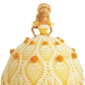 May include: A blonde doll wearing a yellow and white crocheted dress with yellow rose accents. The dress has a full skirt and a fitted bodice. The doll has blue eyes and is posed with her hands on her hips. The dress is a handmade item.