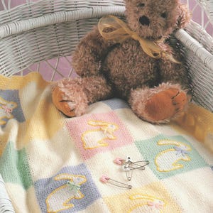 May include: A brown teddy bear with a yellow ribbon sits in a white wicker bassinet. A pastel-coloured baby blanket with embroidered bunnies and safety pins is in the bassinet.