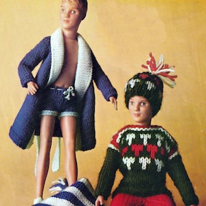 May include: Two dolls are dressed in knitted outfits. One doll wears a blue robe with white trim and matching shorts. The other doll is wearing a green sweater with red and white accents, red pants, and a matching hat. A blue and white striped scarf with fringe is also present.