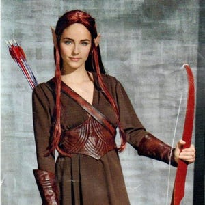 May include: A woman in an elf costume, featuring a brown dress, leather detailing, and tall brown boots. She holds a red bow and arrow. The costume is reminiscent of a fantasy character.