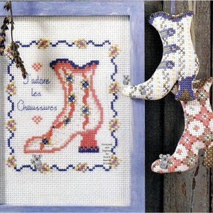 Könnte beinhalten: Ein gerahmtes Kreuzstichbild mit einem Stiefelmotiv, dem Text J'adore les Chaussures und kleinen Koala-Motiven. Rechts hängen zwei weitere dreidimensionale, ausgestopfte Kreuzstich-Stiefelanhänger, die mit floralen und geometrischen Mustern verziert sind.