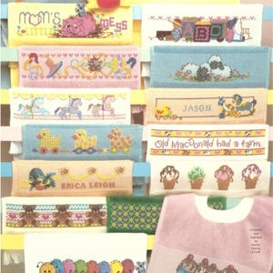 May include: A collection of cross-stitch designs on small towels and bibs. The designs include names, animals, and phrases like "Old MacDonald had a farm." Colors include blue, pink, yellow, and green. A yellow teddy bear is at the top.