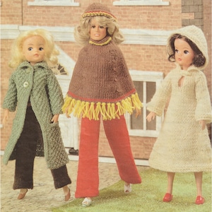 Puede incluir: Tres muñecas vestidas con trajes de punto. Una muñeca lleva un abrigo verde sobre pantalones oscuros. Otra muñeca lleva un poncho marrón con flecos amarillos y pantalones rojos. La tercera muñeca lleva un vestido con capucha color crema.