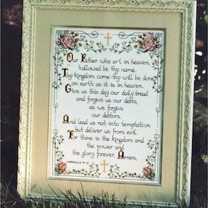 May include: Framed cross-stitch artwork featuring the Lord's Prayer. The text is in black, surrounded by floral and cross motifs in a cream-colored frame. The artwork includes the text "Matthew 6-9-13".